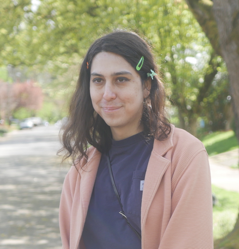 portrait of Abby Castillo. it's a sunny day, and she's smiling in a residential neighborhood with lots of trees in it. she's wearing a blue carhartt shirt, a pink jacket, several hairclips, and some black eyeliner.
